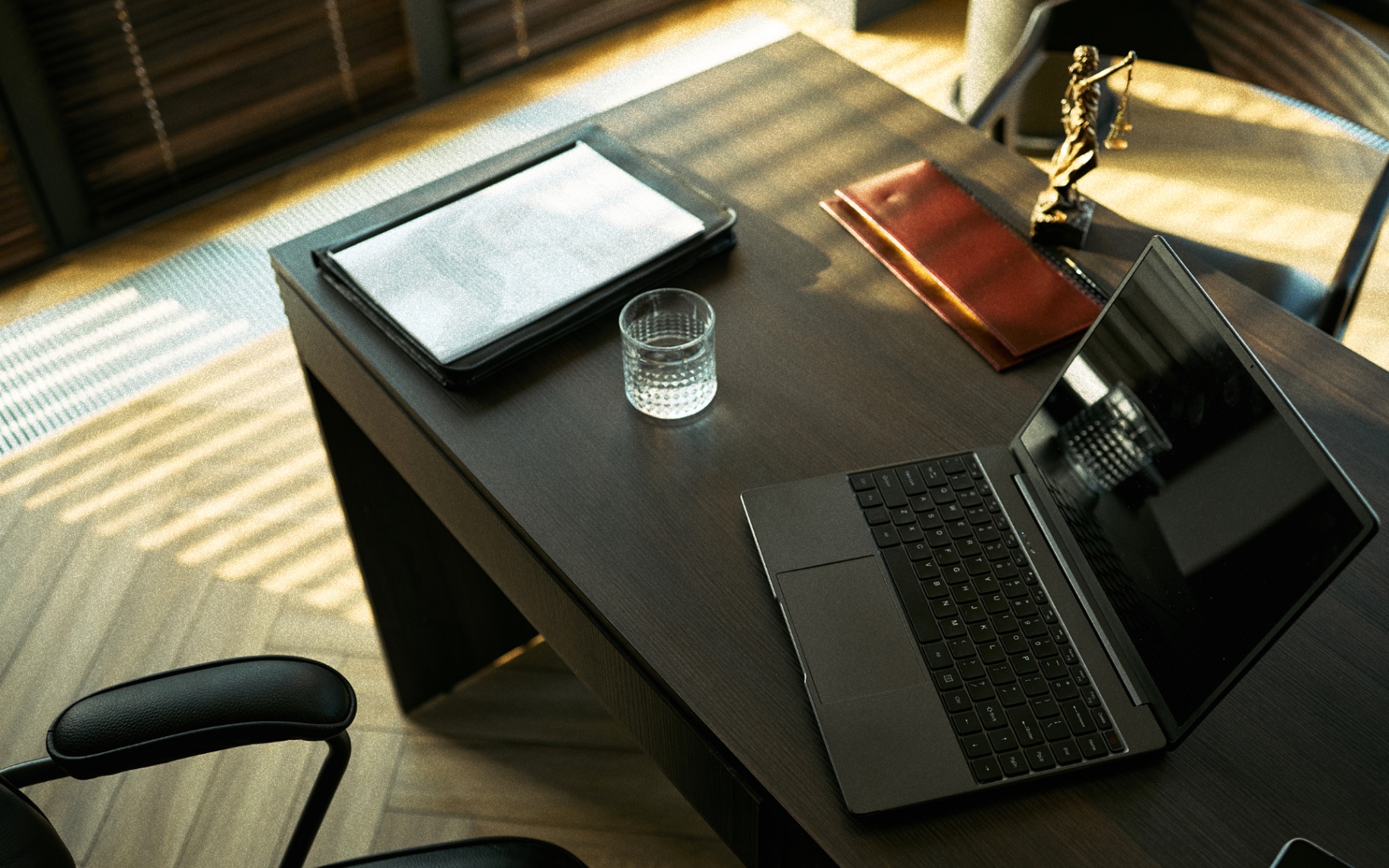 Dunkler, eleganter Schreibtisch mit Laptop, Akten und Justitia-Figur im Sonnenlicht